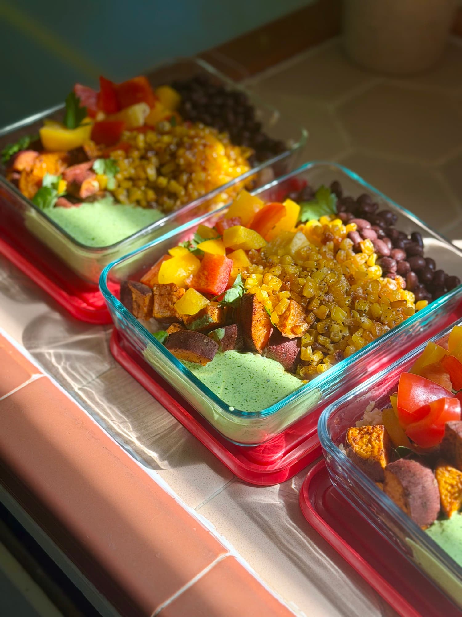 Glass containers of roasted sweet potato, charred corn, black beans and herb sauce on a sunlit counter